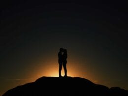 A silhouette of a couple standing on top of a hill at sunset.